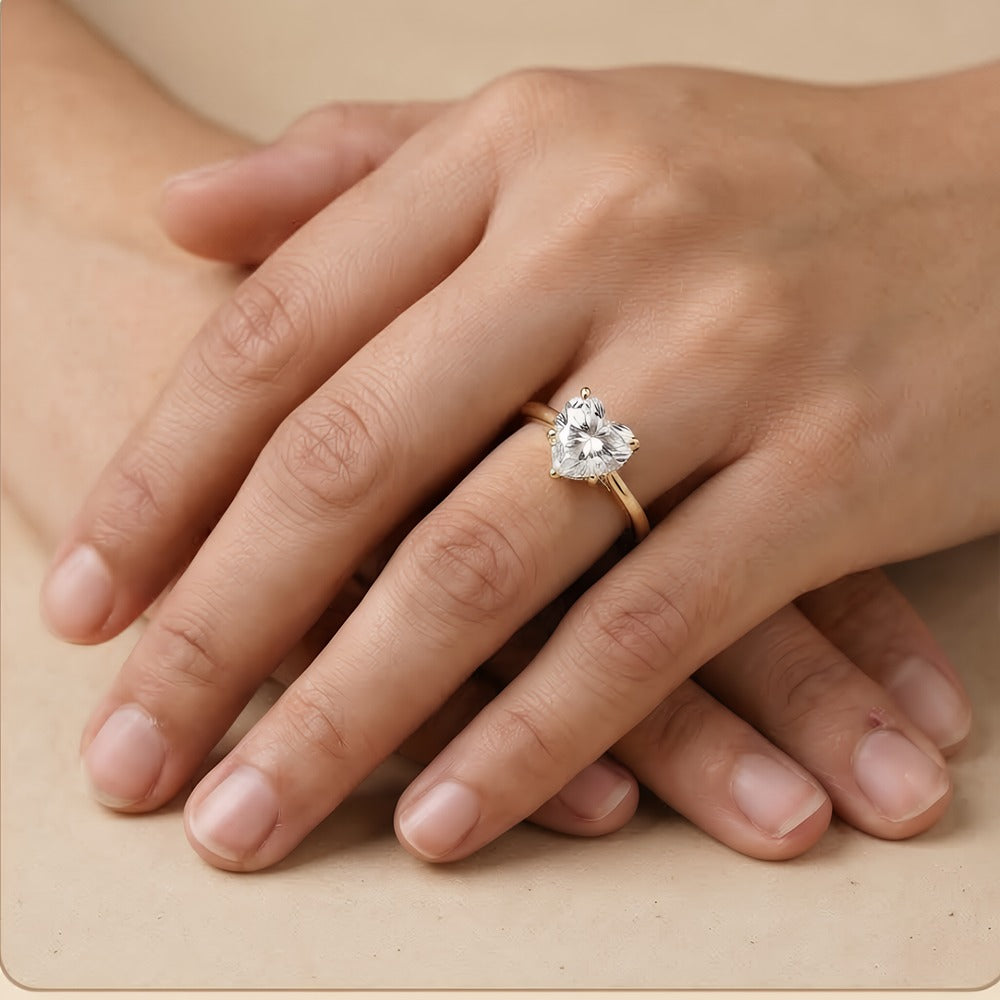A heart-shaped diamond engagement ring on a gold band, shown worn on a person's hand resting on a beige surface.