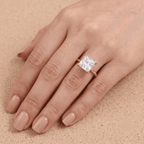 A close-up of a hand wearing a rose gold ring with a square clear gemstone, set against a textured beige background.