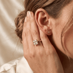 A close-up lifestyle shot of a woman wearing a square-cut diamond engagement ring on her ring finger, with a small gold stud earring on her ear lobe. She is holding her hand near her face against a soft, warm cream satin background.