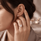 A close-up lifestyle shot of a woman wearing a heart-shaped diamond ring on her finger and a matching heart stud earring, with her hand gently touching her ear.