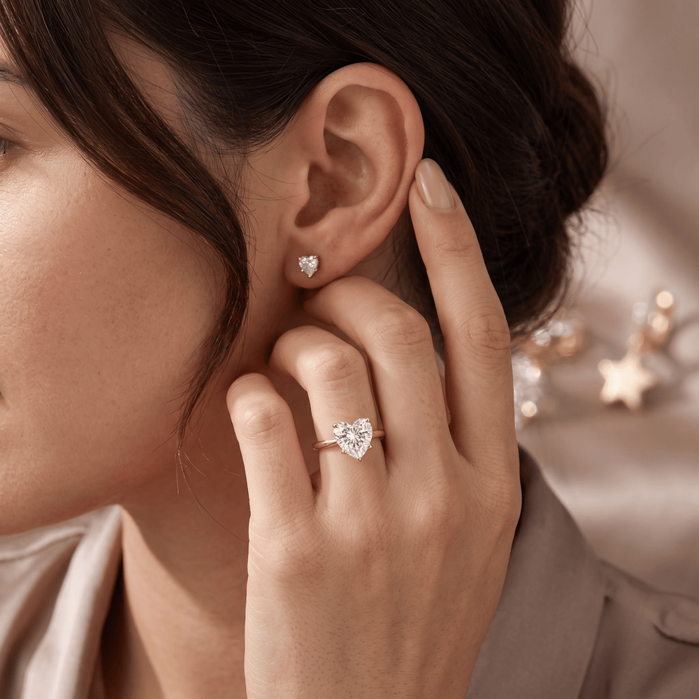 A close-up lifestyle shot of a woman wearing a heart-shaped diamond ring on her finger and a matching heart stud earring, with her hand gently touching her ear.