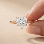 A heart-shaped clear gemstone ring on a gold band, held between two fingers against a soft beige background.