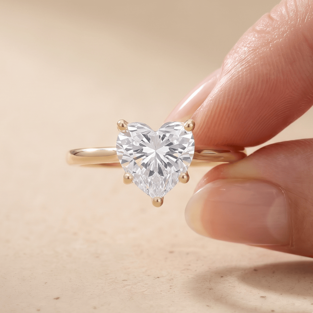 A heart-shaped clear gemstone ring on a gold band, held between two fingers against a soft beige background.