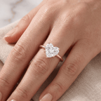 A close-up of a hand wearing a rose gold ring with a heart-shaped clear gemstone, resting on a beige textured fabric.