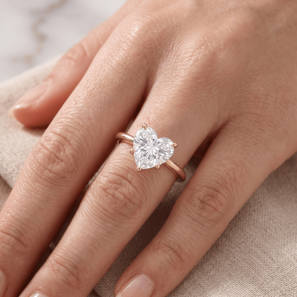 A close-up of a hand wearing a rose gold ring with a heart-shaped clear gemstone, resting on a beige textured fabric.