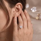 A close-up lifestyle shot of a woman wearing a heart-shaped diamond ring on a rose gold band, with her hand resting on her chin. She also has a small gold hoop in her ear lobe.
