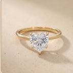 A gold ring featuring a large, heart-shaped clear gemstone in a prong setting, displayed on a light beige textured surface.