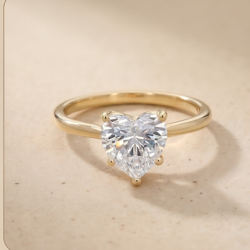 A gold ring featuring a large, heart-shaped clear gemstone in a prong setting, displayed on a light beige textured surface.