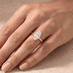 A close-up of a hand wearing a rose gold engagement ring with a marquise-cut diamond. The ring is shown on the ring finger against a neutral, textured beige background.