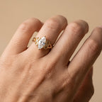 A gold ring with a marquise-cut clear gemstone worn on a person's finger, shown against a soft beige background.