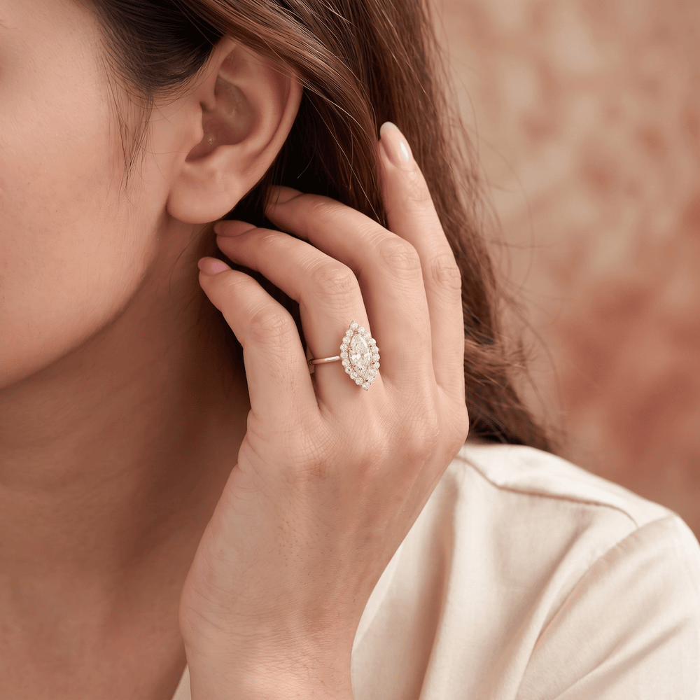 A close-up lifestyle shot of a woman wearing a silver-toned halo ring with a marquise-cut diamond cluster. The ring is shown on her finger, with her hand resting near her ear, set against a soft, warm, textured beige background.
