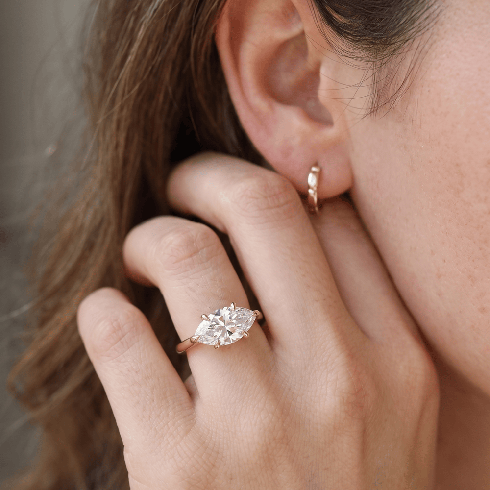 A close-up lifestyle shot of a woman wearing a rose gold ring with a marquise-cut clear gemstone on her ring finger, alongside a matching rose gold hoop earring.