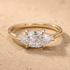 A yellow gold ring featuring a marquise-cut clear gemstone in a four-prong setting, resting on a textured beige sandy surface.