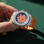 Luxury watch with orange strap and blue face held by a hand on a green background