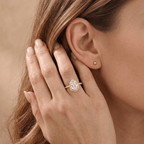 A close-up lifestyle shot of a woman's hand and ear wearing a silver halo ring with a clear teardrop-shaped center stone and smaller surrounding stones. She also wears a small matching stud earring.