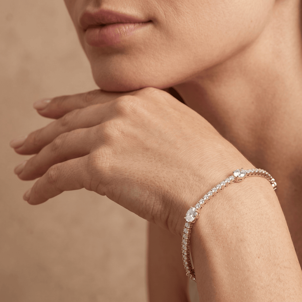 A close-up lifestyle shot of a model wearing a delicate diamond tennis bracelet on her wrist, with her hand resting near her chin against a warm beige background.
