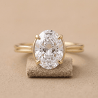An oval-cut clear gemstone ring set in a yellow gold band, displayed on a small textured beige stand against a warm beige background.