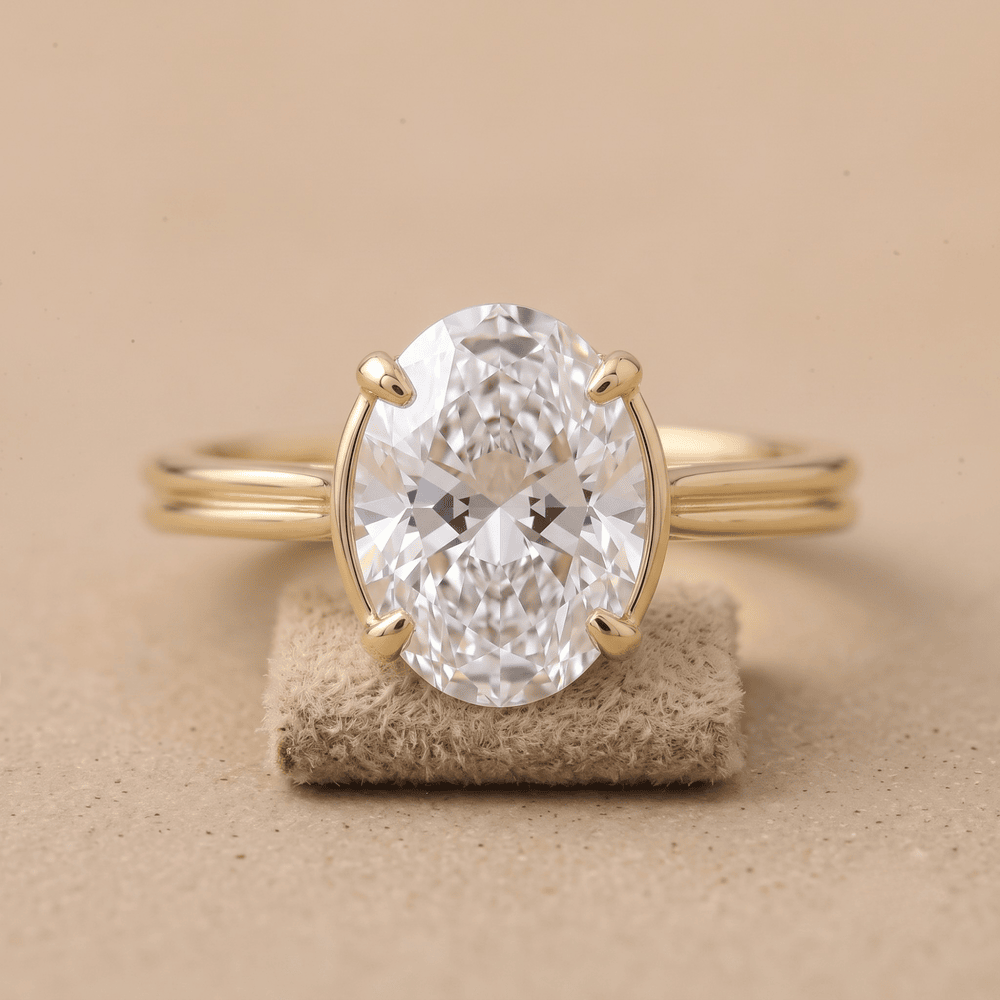 An oval-cut clear gemstone ring set in a yellow gold band, displayed on a small textured beige stand against a warm beige background.