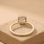 A yellow gold ring with a round clear gemstone in a prong setting, displayed on a textured beige stone block against a warm beige background.