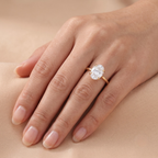 A close-up of a hand wearing a yellow gold ring with an oval-cut clear gemstone, set against a soft beige skin-toned background.