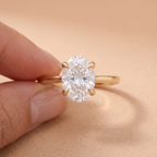 A close-up of a hand holding a gold ring with an oval-cut clear gemstone against a soft beige background.