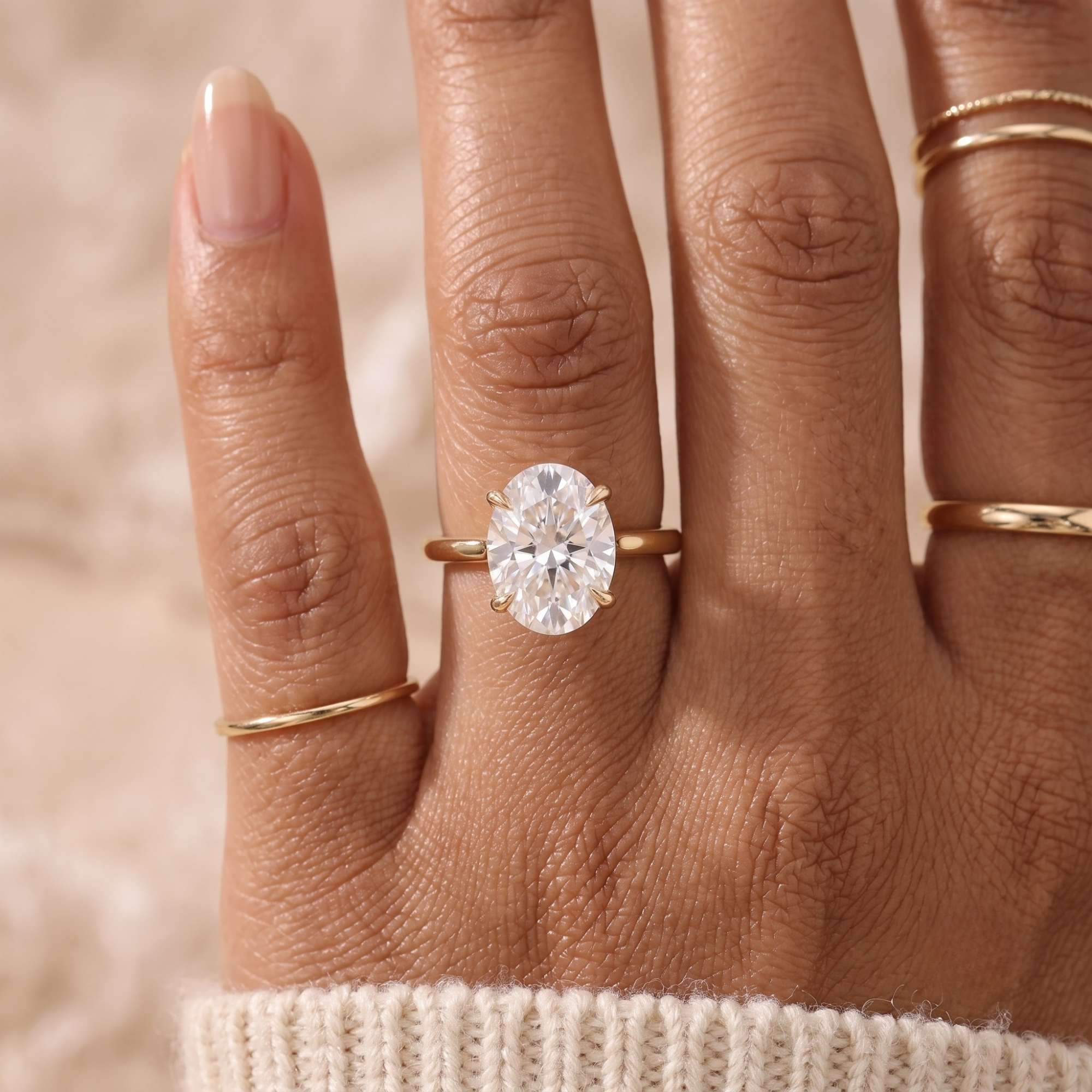 A close-up of a hand wearing a gold ring with a large oval clear gemstone, along with two thin gold bands on other fingers, set against a soft beige background.