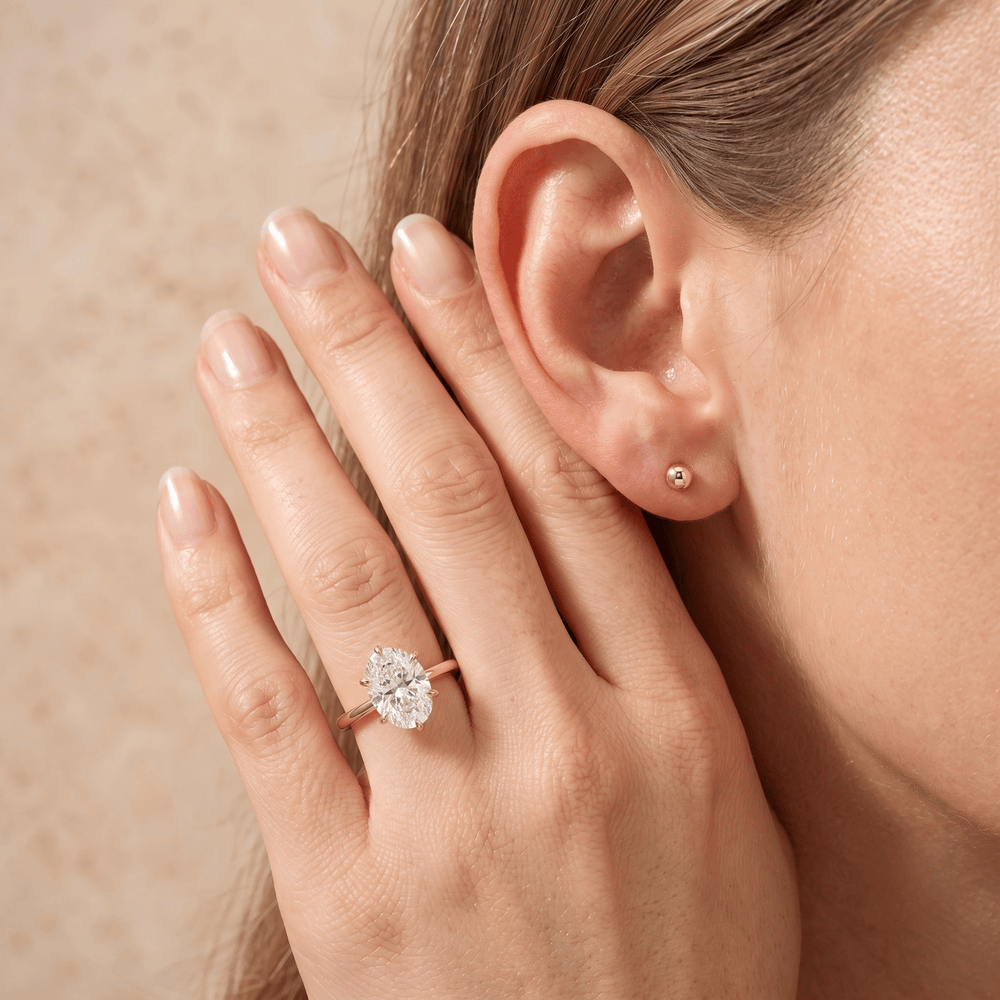 A close-up lifestyle shot of a woman's hand and ear, showcasing a rose gold ring with a marquise-cut clear gemstone on her ring finger, alongside a matching rose gold stud earring.