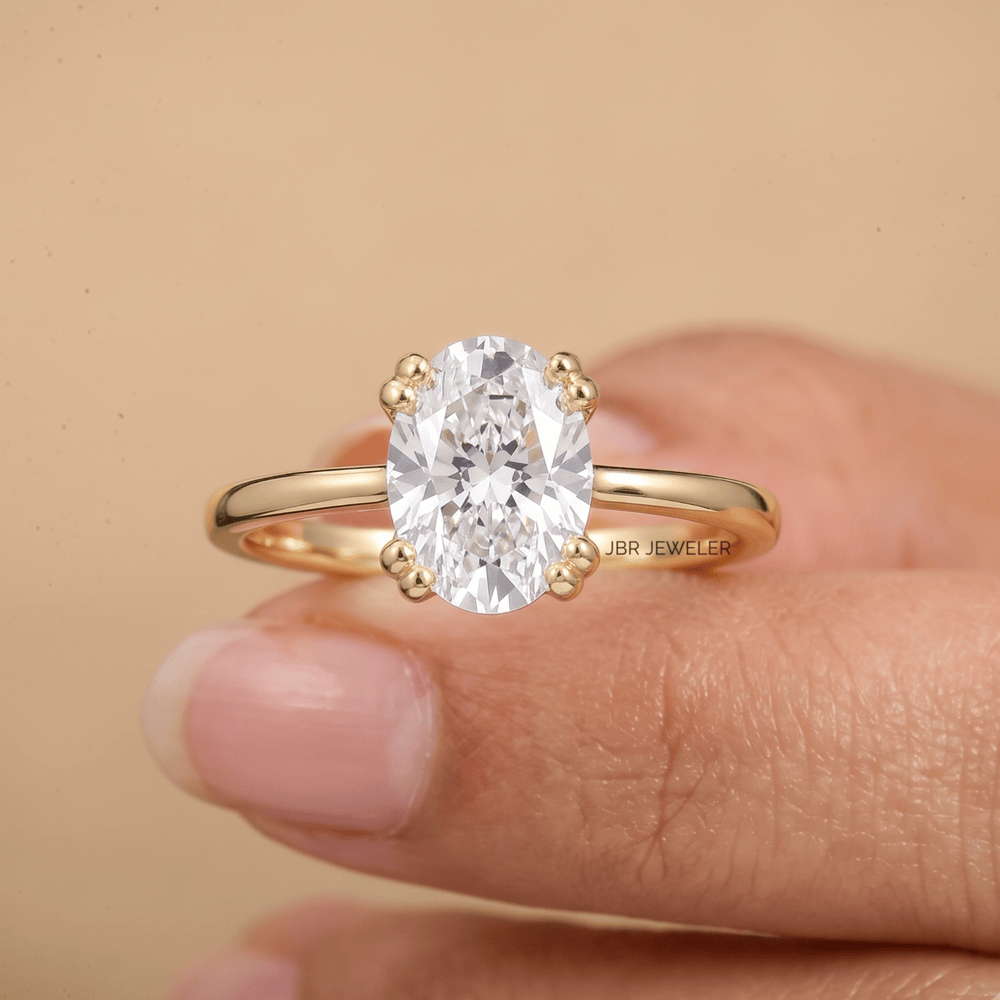 A yellow gold ring with a large oval-cut clear gemstone in a four-prong setting, held on a person's fingers against a warm beige background. The brand name 'JBR JEWELER' is visible on the inner band.