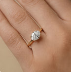 An oval-cut diamond engagement ring with a yellow gold band, shown worn on a person's finger against a soft beige background.