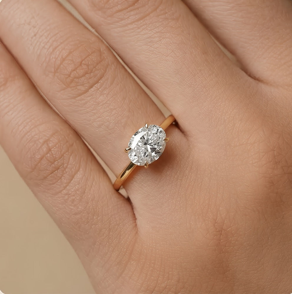 An oval-cut diamond engagement ring with a yellow gold band, shown worn on a person's finger against a soft beige background.