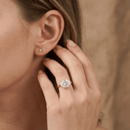 A close-up lifestyle shot of a woman's hand wearing a rose gold ring with a large oval-cut clear gemstone, and a matching small stud earring in her other ear. The background is a soft, warm beige fabric.