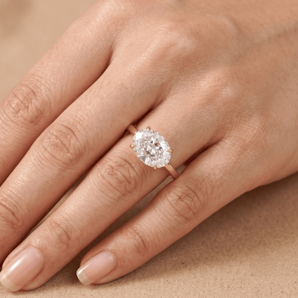 A close-up of a hand wearing a rose gold ring with a large oval-cut clear gemstone, set against a soft beige background.