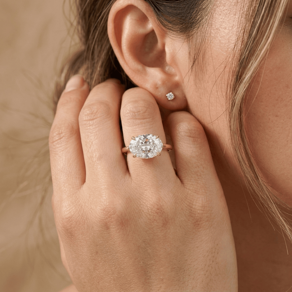 A close-up lifestyle shot of a woman wearing a rose gold ring with a round brilliant-cut diamond and a matching small diamond stud earring. The ring is shown on her ring finger, with her hand resting near her ear against a soft, warm beige background.