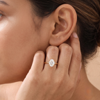 A close-up lifestyle shot of a woman wearing a gold ring with a round-cut clear gemstone on her finger, with her hand resting near her ear.