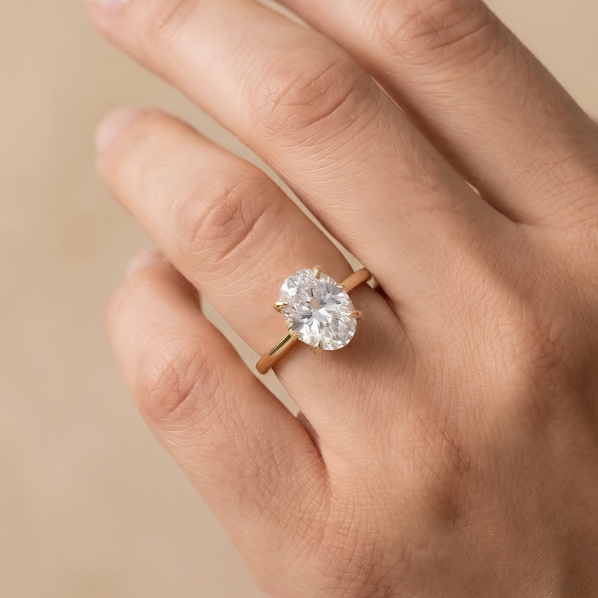 A close-up of a hand wearing a classic solitaire engagement ring with a round brilliant-cut diamond on a rose gold band, set against a soft beige background.