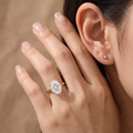 A close-up lifestyle shot of a woman's hand wearing a rose gold engagement ring with a round-cut clear gemstone. The hand is resting against her ear, which has a small gold stud earring, against a soft, warm brown background.