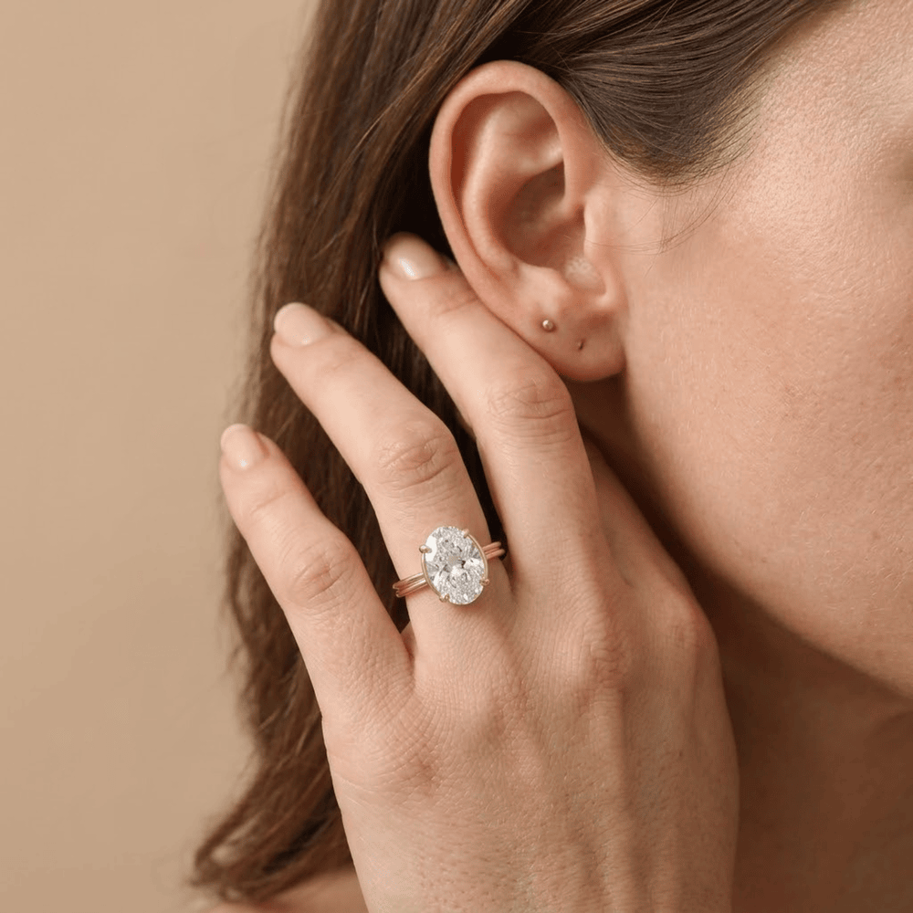 A close-up lifestyle shot of a woman wearing a rose gold ring with a large clear oval gemstone cluster, shown on her finger against a warm beige background.