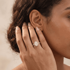 A close-up lifestyle shot of a woman wearing a silver halo ring with a round-cut clear center stone on her ring finger, with a matching small stud earring on her ear lobe.