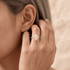 A close-up lifestyle shot of a woman wearing a rose gold engagement ring with a round-cut diamond on her ring finger. The warm, sepia-toned image highlights the delicate band and the sparkling diamond, with her ear and hair visible in the background.