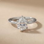 A silver solitaire engagement ring with a pear-cut diamond center stone, set against a warm beige background.