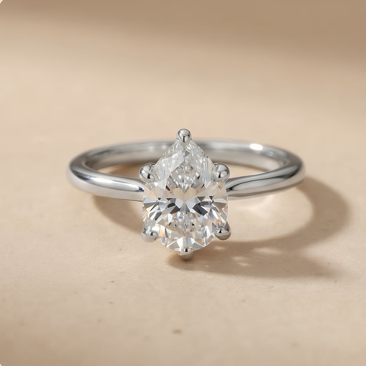 A silver solitaire engagement ring with a pear-cut diamond center stone, set against a warm beige background.