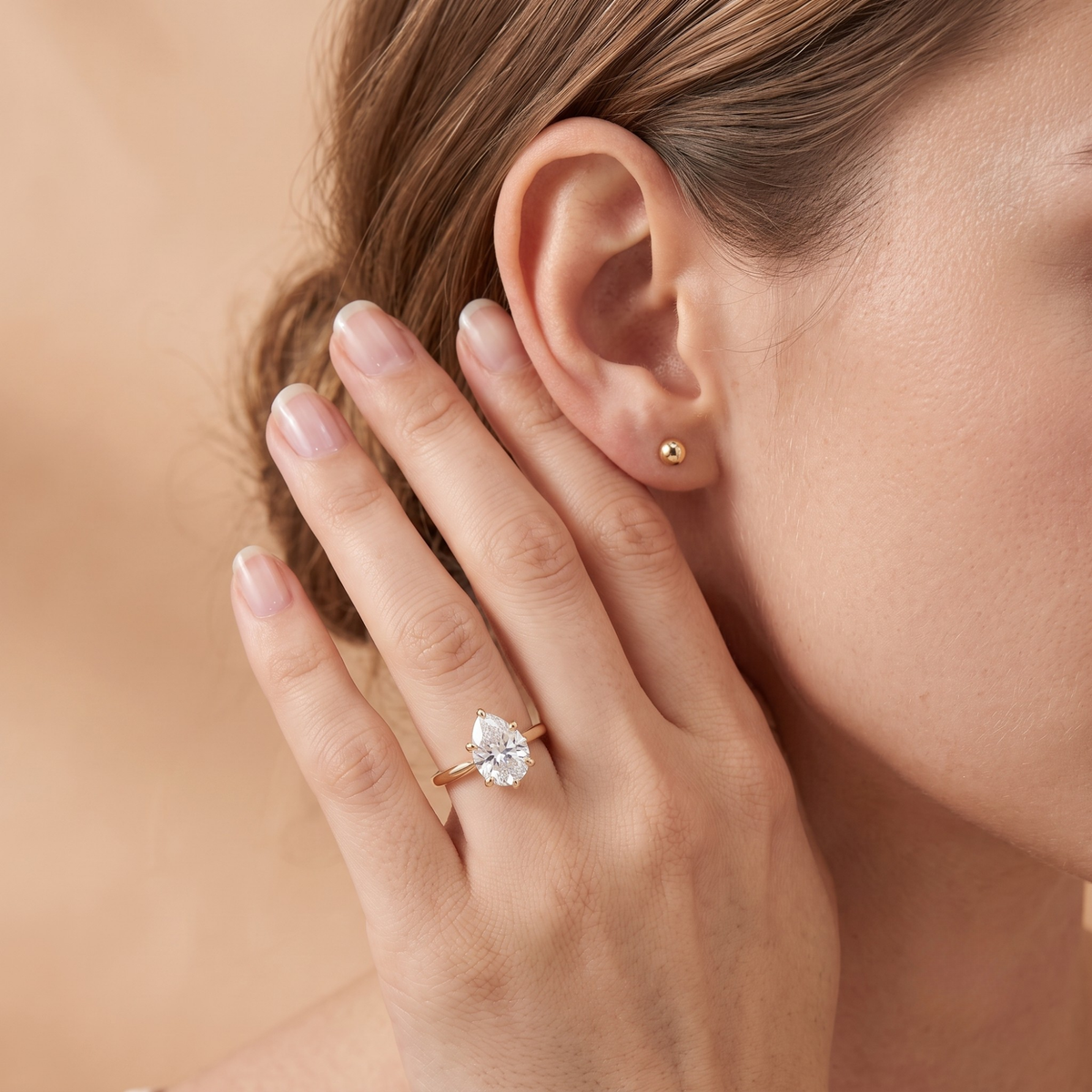 A close-up lifestyle shot of a woman wearing a pear-cut diamond ring on a rose gold band and a small gold stud earring. Her hand is gently resting against her ear, set against a soft, warm beige background.