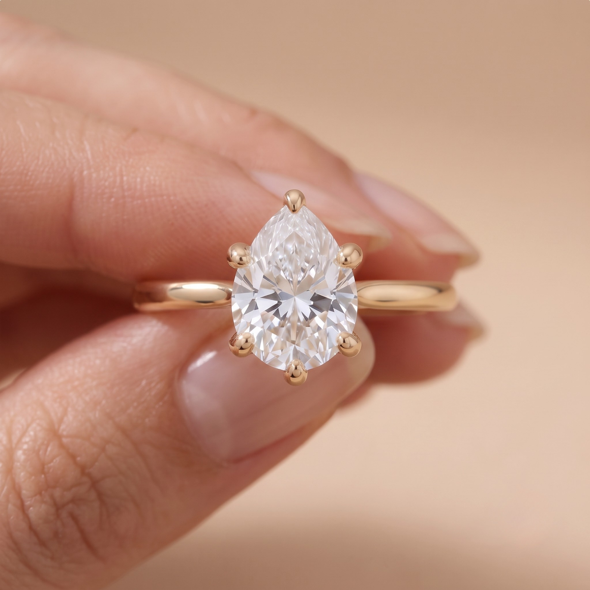 A close-up of a hand holding a gold ring with a large pear-cut clear gemstone in a prong setting, against a soft beige background.