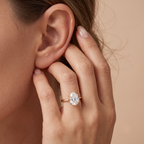 A close-up lifestyle shot of a woman's hand and ear wearing a rose gold ring with a pear-cut clear gemstone on her ring finger, set against a soft beige background.