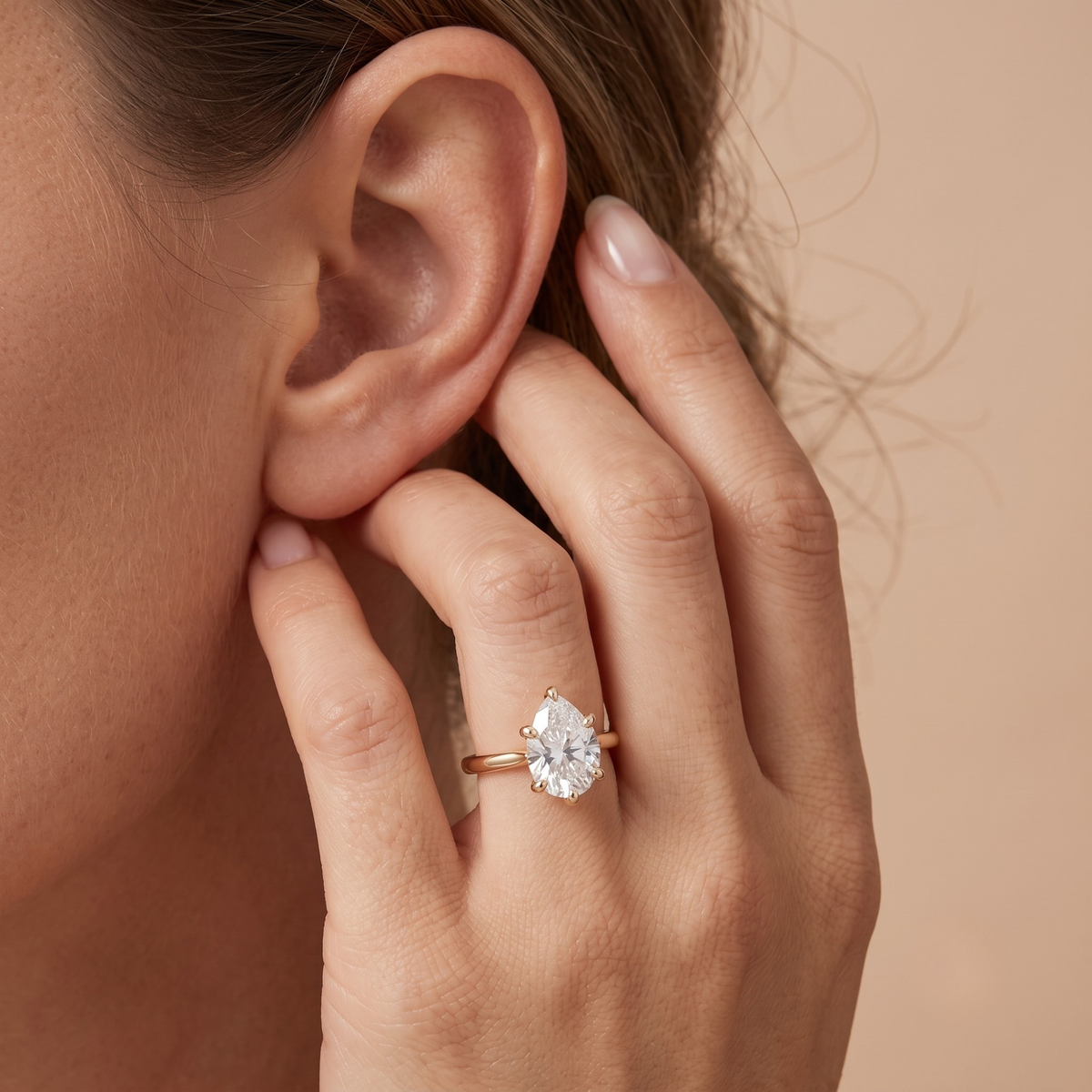 A close-up lifestyle shot of a woman's hand and ear wearing a rose gold ring with a pear-cut clear gemstone on her ring finger, set against a soft beige background.