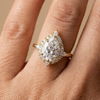 A close-up of a hand wearing a pear-shaped diamond engagement ring with a halo of smaller diamonds in a yellow gold setting.