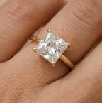 A gold ring with a large square princess-cut clear gemstone in a four-prong setting, shown worn on a hand.