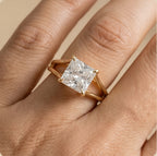 A close-up of a hand wearing a gold ring with a princess-cut clear gemstone. The ring features a four-prong setting on a split shank band, shown against a warm beige skin and background.