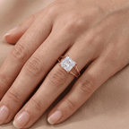 A close-up of a hand wearing a rose gold ring with a square princess-cut diamond. The ring is shown on the ring finger against a soft beige background.