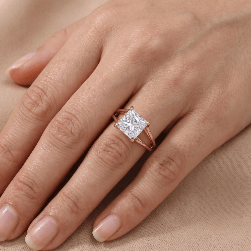 A close-up of a hand wearing a rose gold ring with a square princess-cut diamond. The ring is shown on the ring finger against a soft beige background.